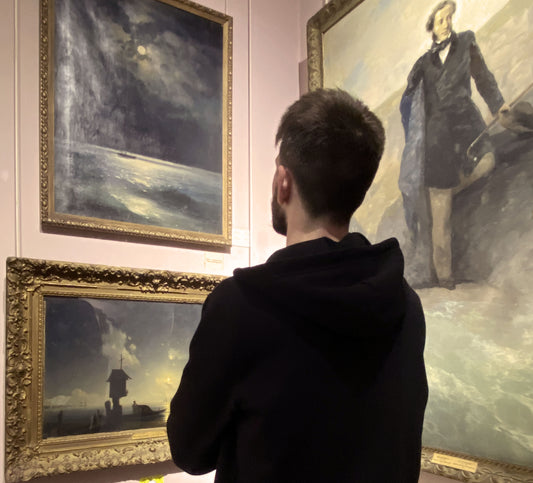 Man in black hoodie before ornate gold-framed paintings in restoration display