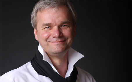 A man with gray hair wearing a white collared shirt and a black scarf.