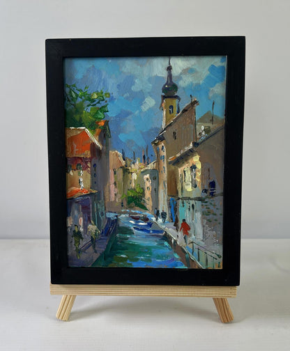 Black-framed oil painting of sunlit canal by Yuriy Suprunchuk on easel