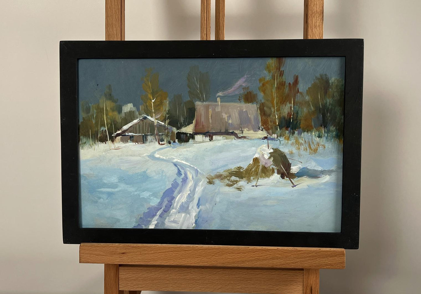 A framed winter landscape painting with snow-covered cottages and trees, enclosed in a simple black frame.