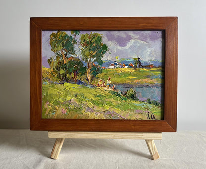 A rustic wooden-framed oil painting depicting a vibrant pastoral scene with lush green trees, rolling fields, and distant figures under a soft purple sky.