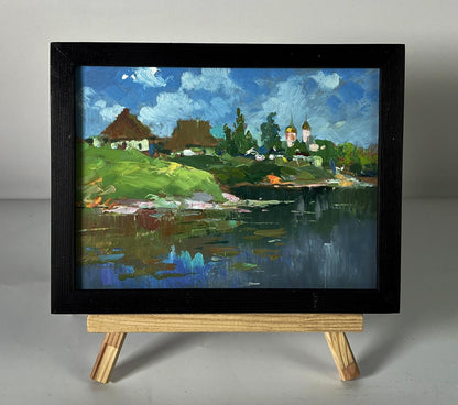 Oil painting of river near village by Yuriy Suprunchuk in black frame on easel