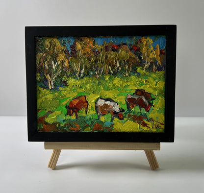 Vibrant oil painting of cows grazing in meadow by Oksana Ivanyuk on wooden easel