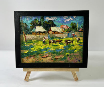 Vibrant impressionist oil painting of cows grazing outside village by Oksana Ivanyuk on easel