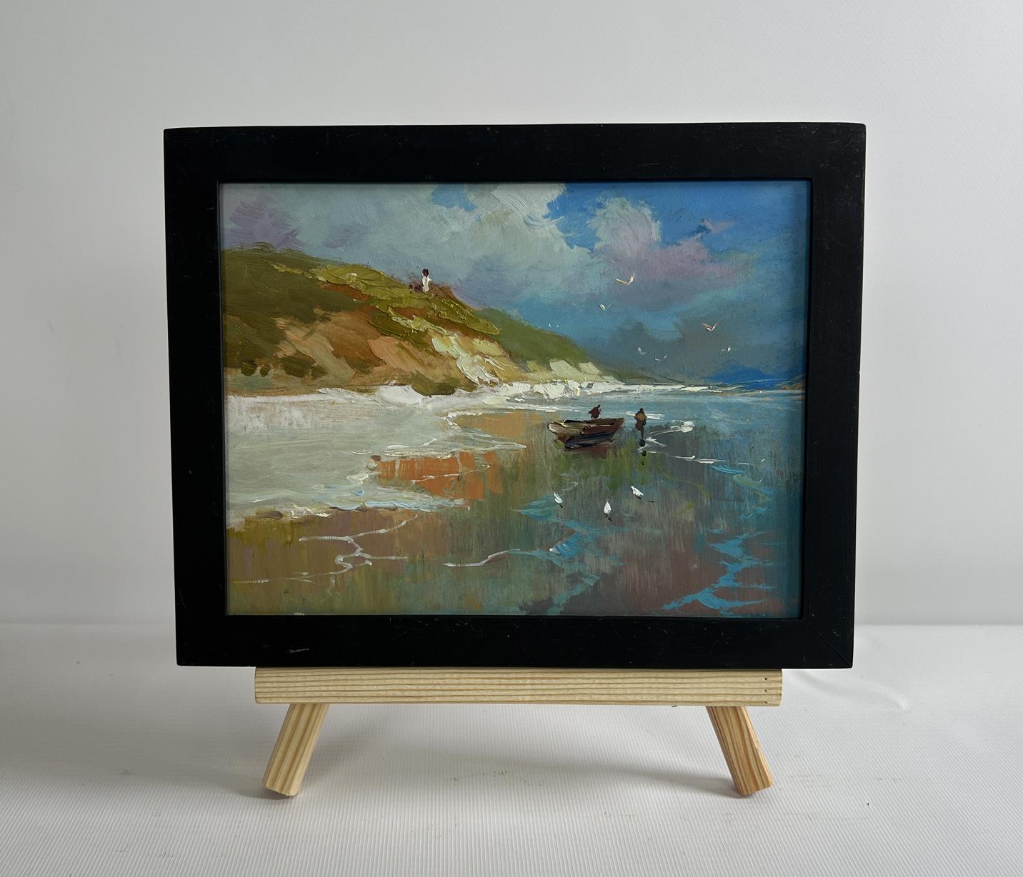 Black-framed miniature painting of a coastal scene with boats and cliffs, displayed on a wooden easel.