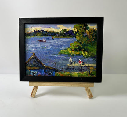 Black-framed oil painting of relaxing evening river scene by Oksana Ivanyuk on wooden easel