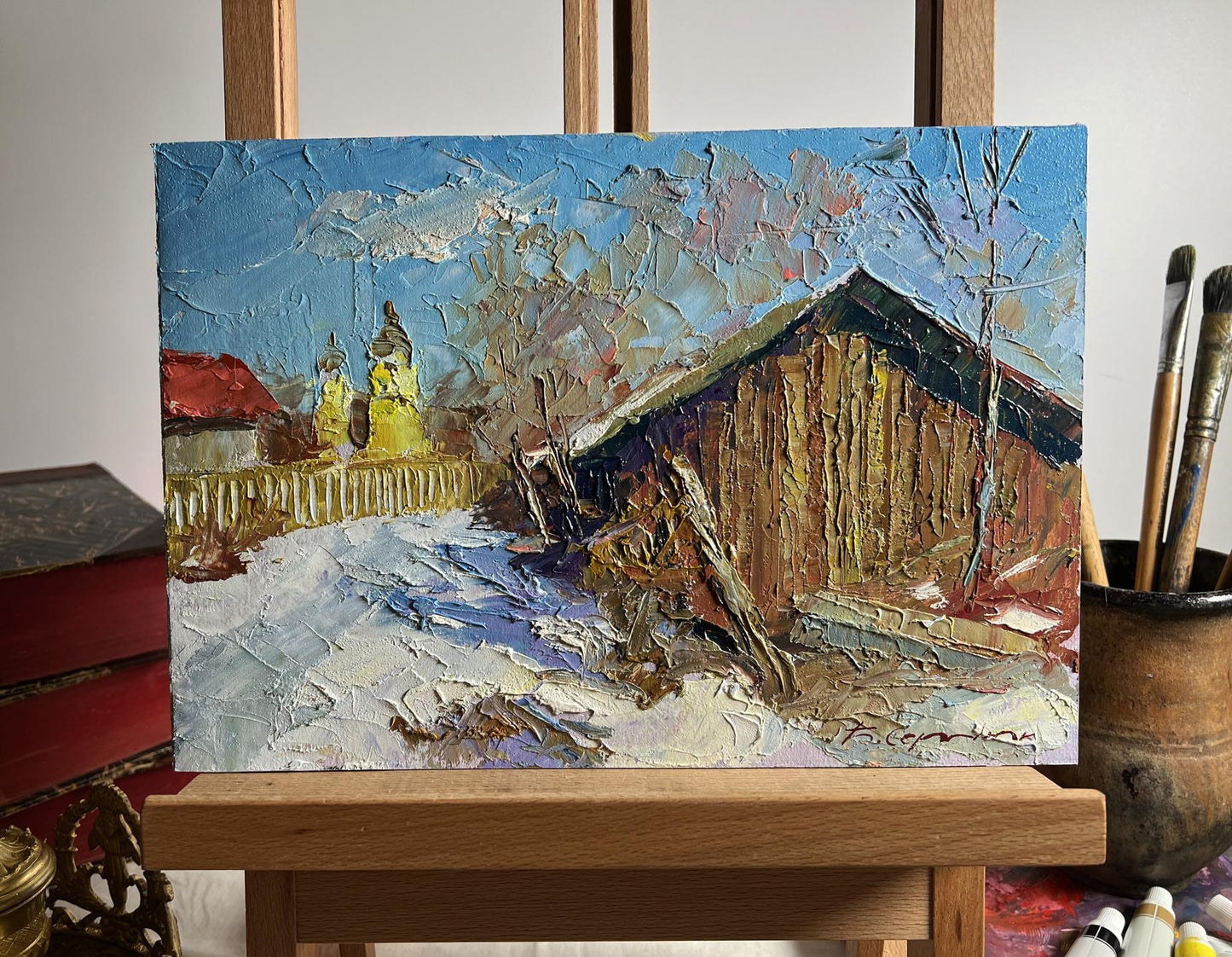 A textured, impressionistic oil painting on canvas depicting a snowy rural scene with a wooden barn and distant church spires under a bright blue sky.