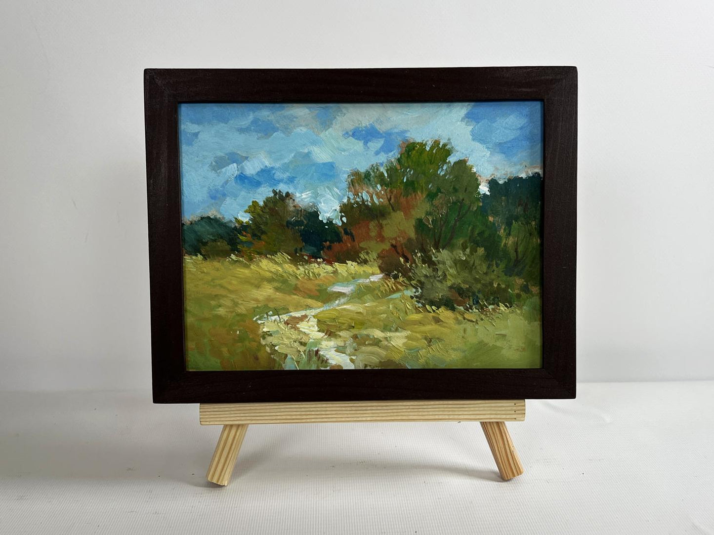 A framed oil painting of a pastoral landscape with vibrant green trees and a winding stream under a blue sky, enclosed in a dark wooden frame.