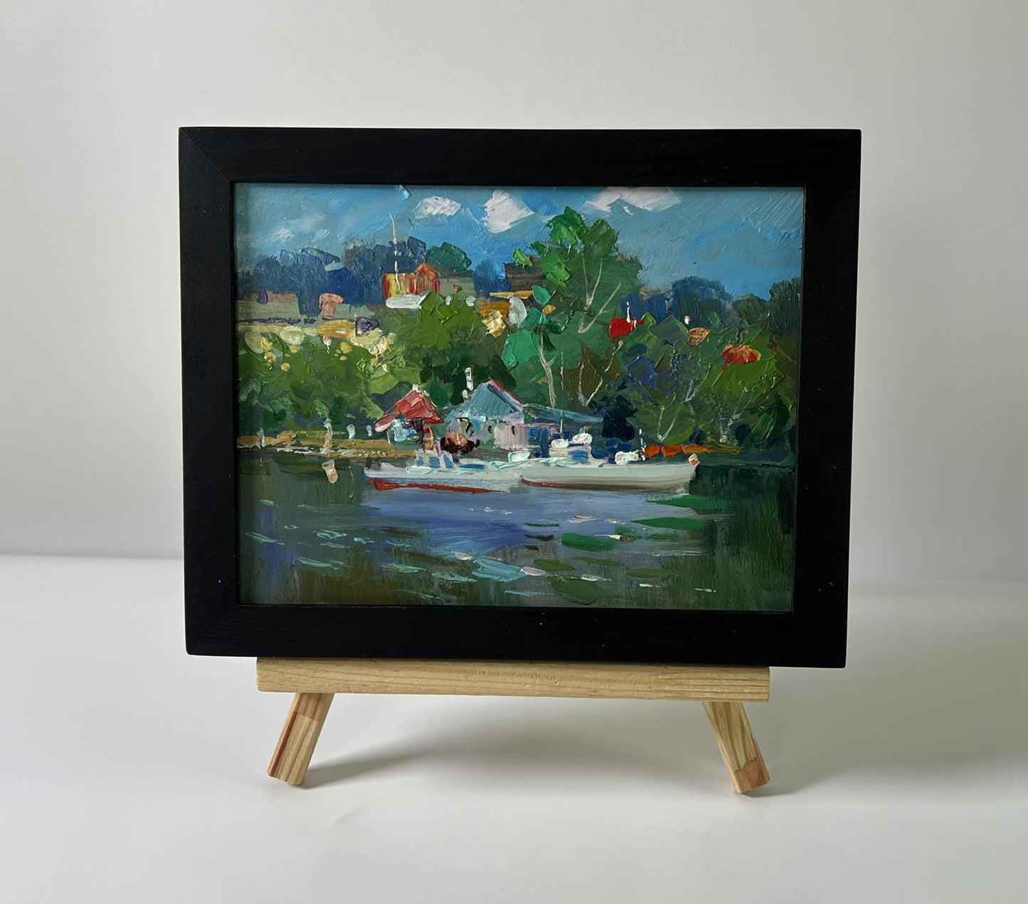 Black-framed oil painting of Yuriy Suprunchuk lakeside village with boats