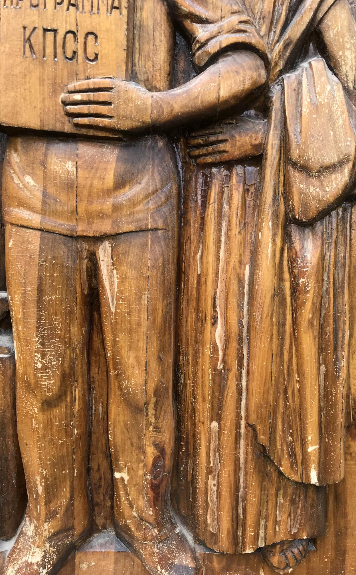 Detailed wooden carving of two figures in traditional attire holding Cyrillic plaque, social realism devotion to communism