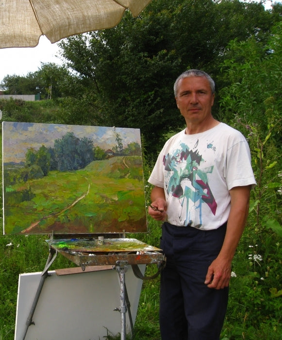 A man in a white t-shirt with a colorful anime-style graphic stands outdoors beside an easel holding a landscape painting.