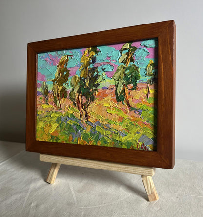A vibrant, textured oil painting of trees and landscape in bold greens, yellows, pinks, and blues, encased in a rich brown wooden frame.