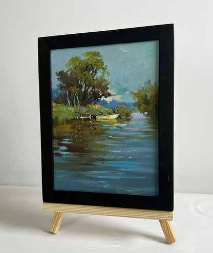 Black-framed oil painting of serene river with boat by Yuriy Suprunchuk on easel