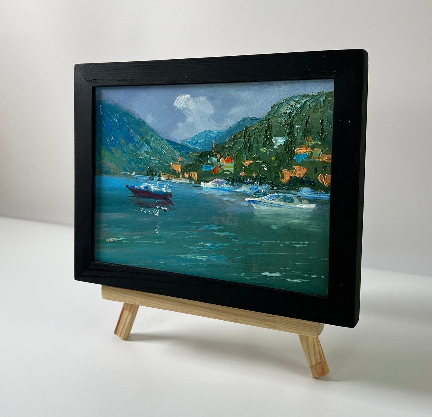 Small black-framed oil painting of serene lakeside boats and mountains by Yuriy Suprunchuk on wooden easel