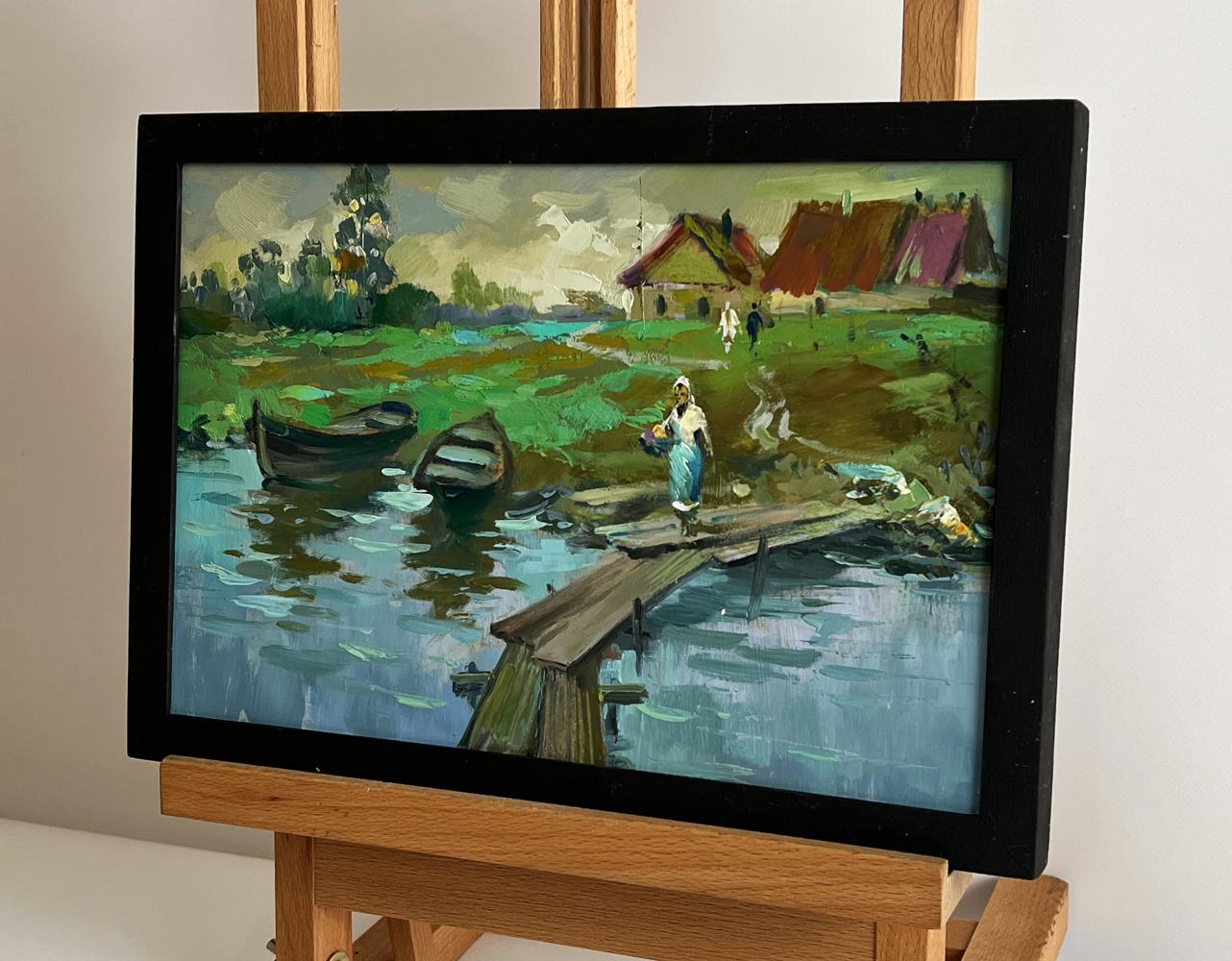 Black-framed oil painting by Yuriy Suprunchuk of village river with boats and bridge
