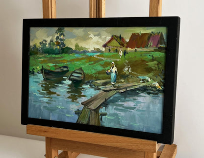Black-framed oil painting by Yuriy Suprunchuk of village river with boats and bridge