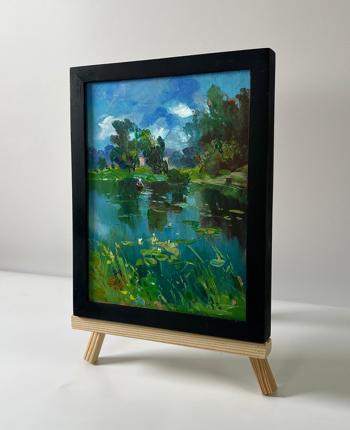 Black-framed oil painting of serene forest river lake with lily pads on easel