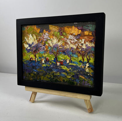 Vibrant oil painting of blossoming orchard by Oksana Ivanyuk in black frame on easel