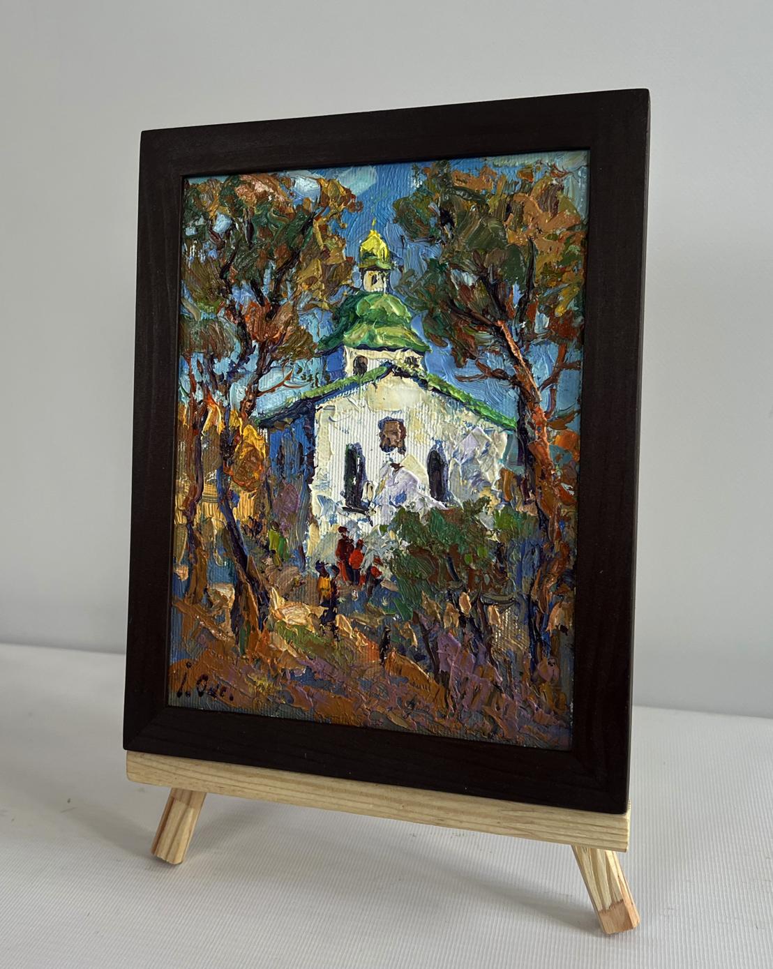A framed oil painting depicting a white church with a green dome nestled among autumn trees, encased in a dark brown wooden frame.