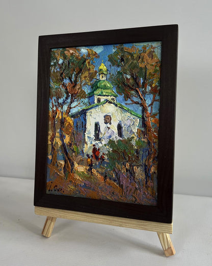 A framed oil painting depicting a white church with a green dome nestled among autumn trees, encased in a dark brown wooden frame.