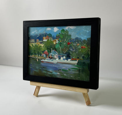 Black-framed oil painting of village boats on ferry by Yuriy Suprunchuk on wooden easel
