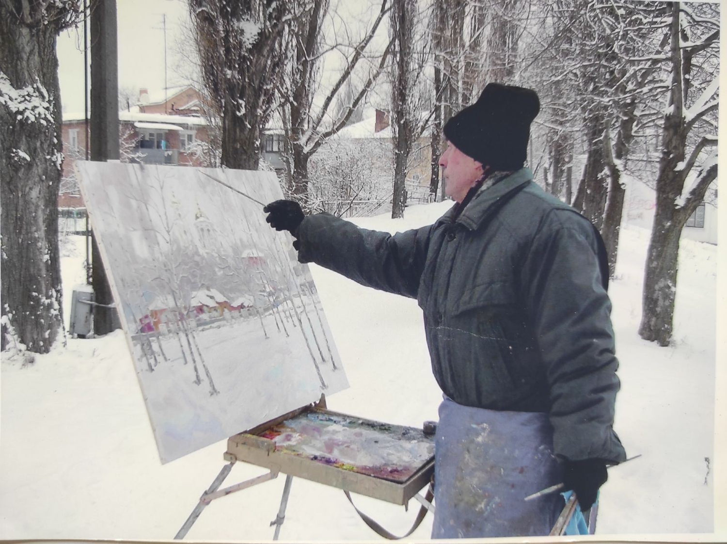 A painter in a black beanie and dark coat stands outdoors, holding a paintbrush to an easel-mounted canvas depicting a snowy landscape with bare trees.