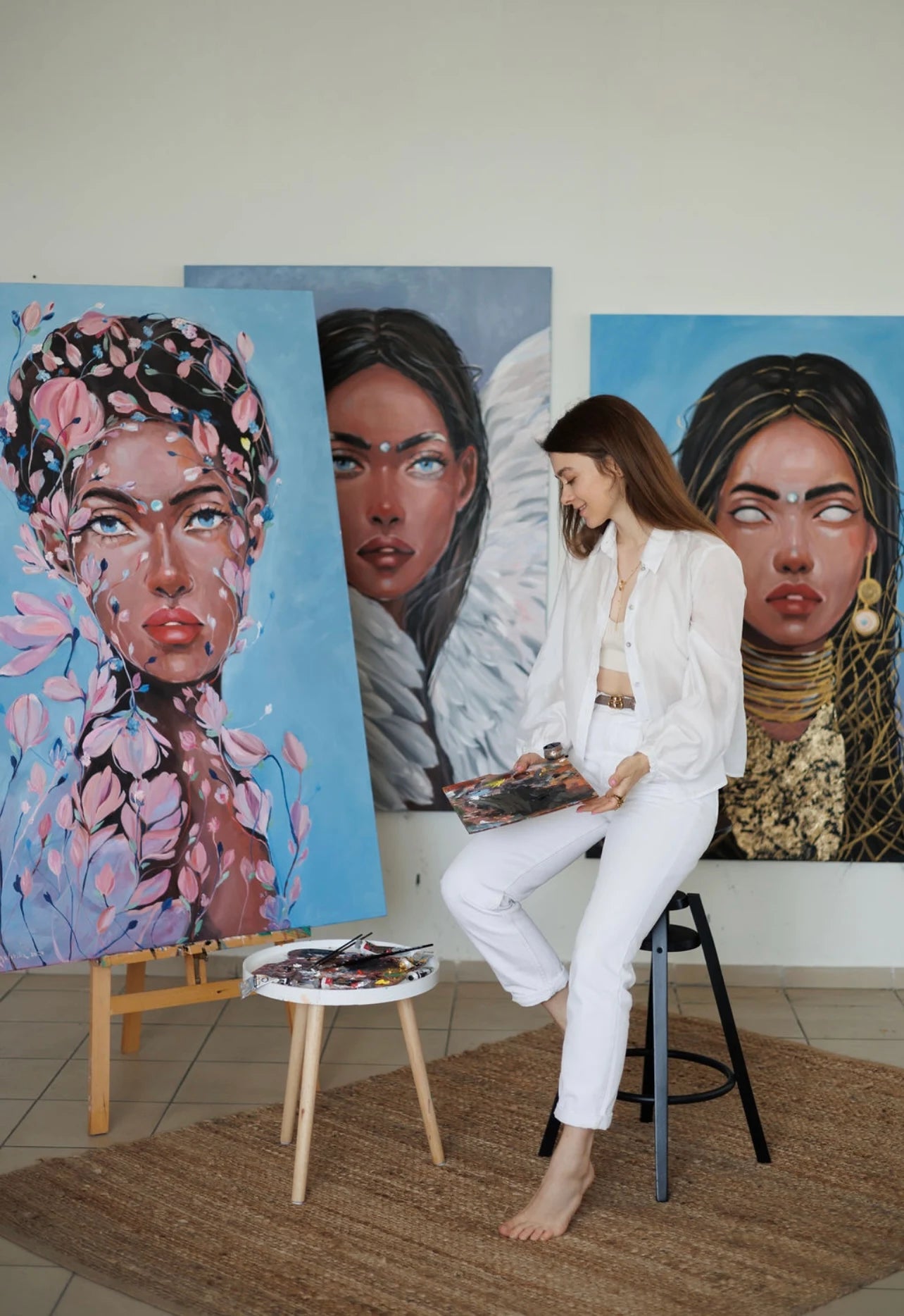 A woman in a white blouse and pants sits barefoot on a black stool, holding an open sketchbook.