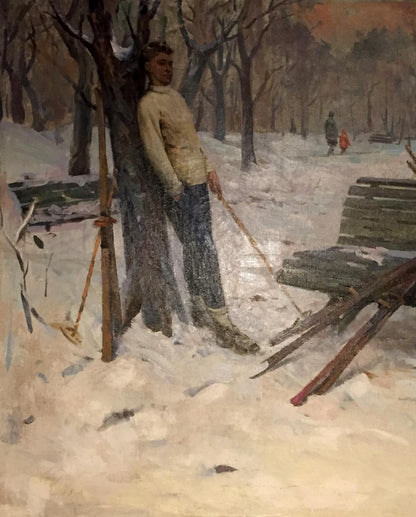 A man in a beige sweater and blue jeans stands leaning against a tree, holding ski poles with snow-covered ground around him.